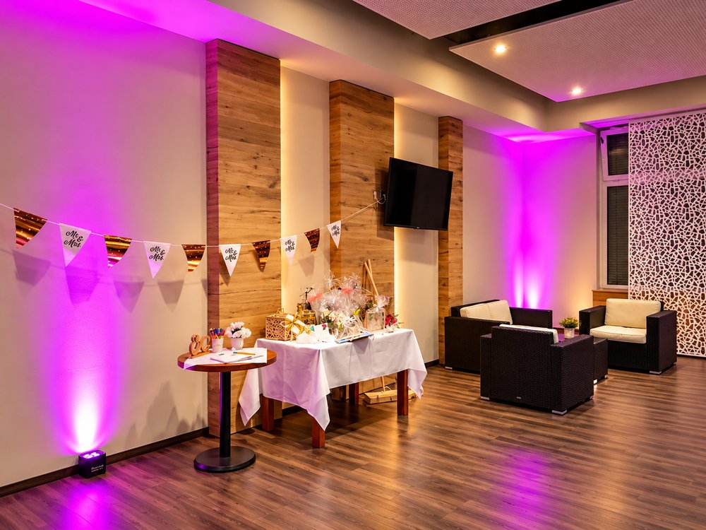 Festsaal mit magentafarbenen Uplights an Wänden und Holzsäulen, Geschenktisch, Wimpelkette und Lounge-Sesseln