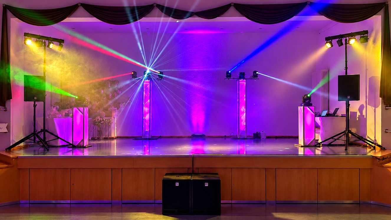 Festsaal-Bühne mit Moving Heads auf Trusstowern, LED-Lichtanlagen und Lautsprechern auf Stativen, farbigen Lichtstrahlen und zwei Subwoofern vor der Bühne (KI-bearbeitet)