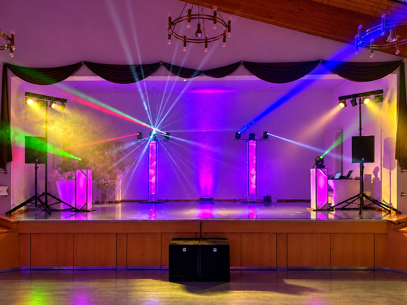 Festsaal-Bühne mit Moving Heads auf Trusstowern, LED-Lichtanlagen und Lautsprechern auf Stativen, farbigen Lichtstrahlen und zwei Subwoofern vor der Bühne (KI-bearbeitet)