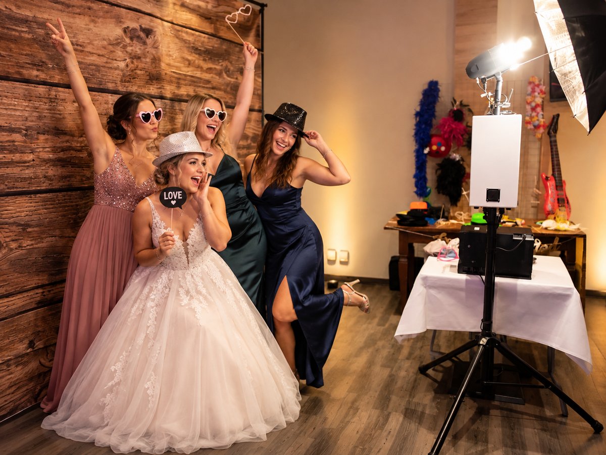 Braut und Freundinnen posieren mit Herzbrillen, Hüten und LOVE-Schild vor der TF-Event-Fotobox mit Studioblitz und Requisitentisch (Personen KI-generiert)