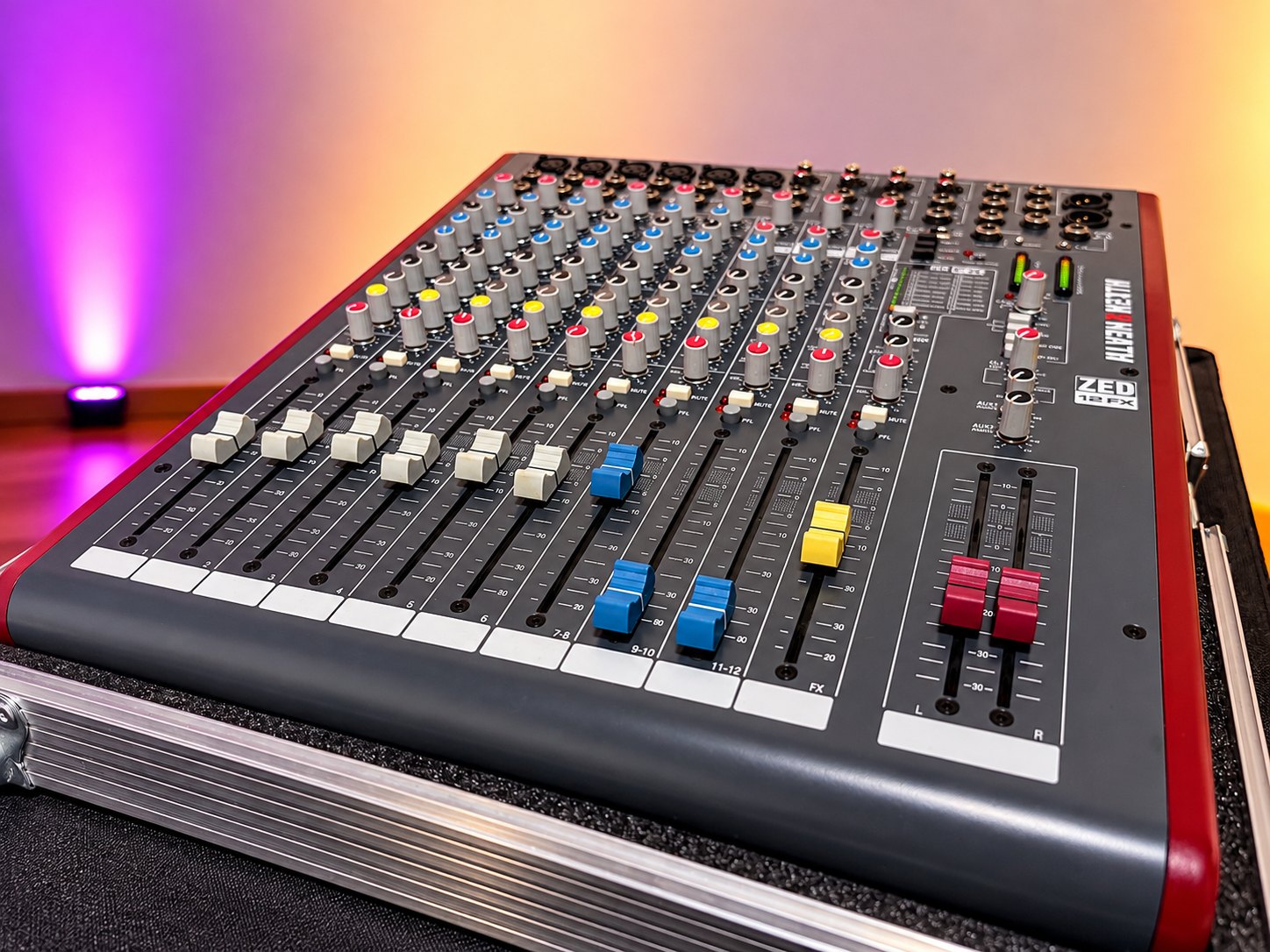 Mischpult Allen & Heath ZED-12FX mit farbigen Reglern und Fadern auf einem Transportcase, im Hintergrund ein magentafarbenes Uplight (Hintergrund KI-generiert)